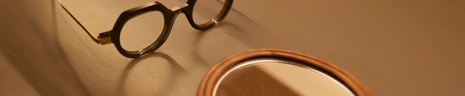 Photo bannière de la page Connivence Opticiens, un epaire de lunettes de marque posées sur une table en bois devant un miroir